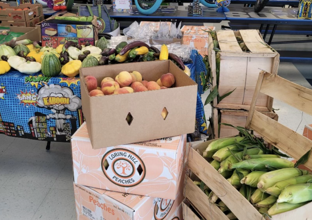 A box of open peaches sits on top of two unopened "Loring Hill Peaches" boxes. Beside the peach boxes is a crate of ears of corn with their husks intact. In the background is a table with a blue tablecloth covered with an assortment of vegetables.