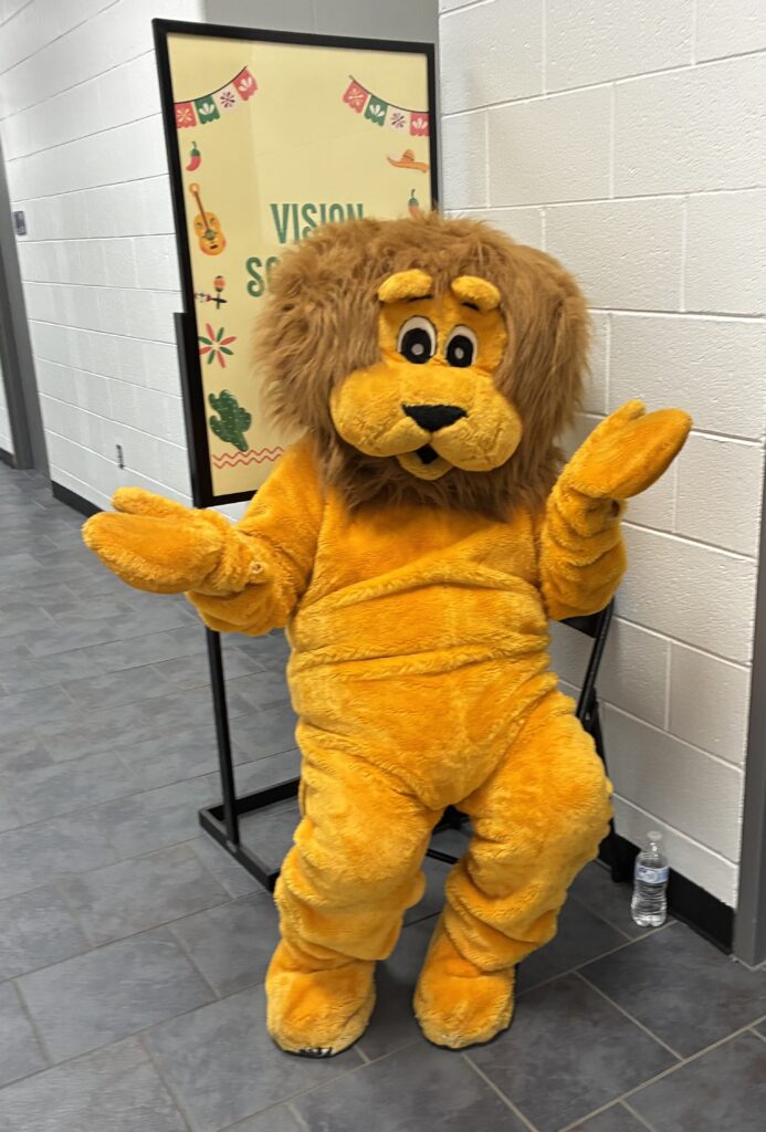 A person in a fluffy lion costume is seated against a white wall, looking playful with shrugged “I don’t know” hands. A colorful sign is visible behind them.