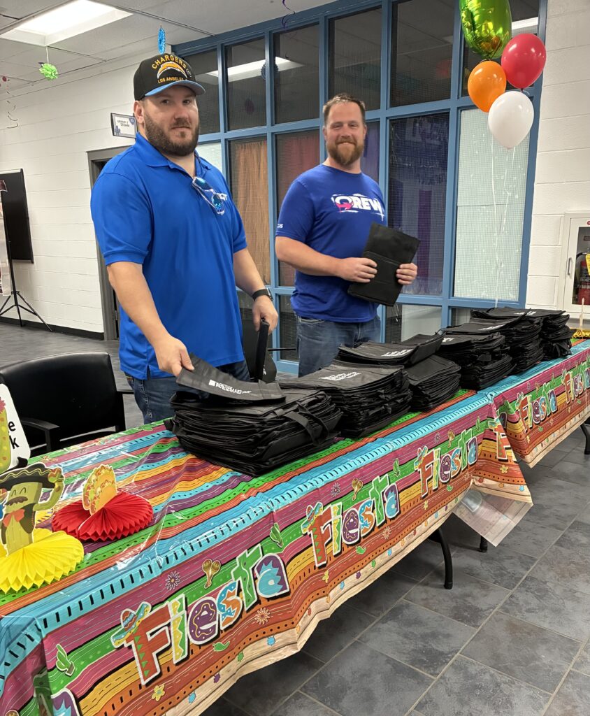 Two men wearing blue shirts stand behind a festive table with "Fiesta" themed decorations, handing out black bags. Bright balloons adorn the scene.