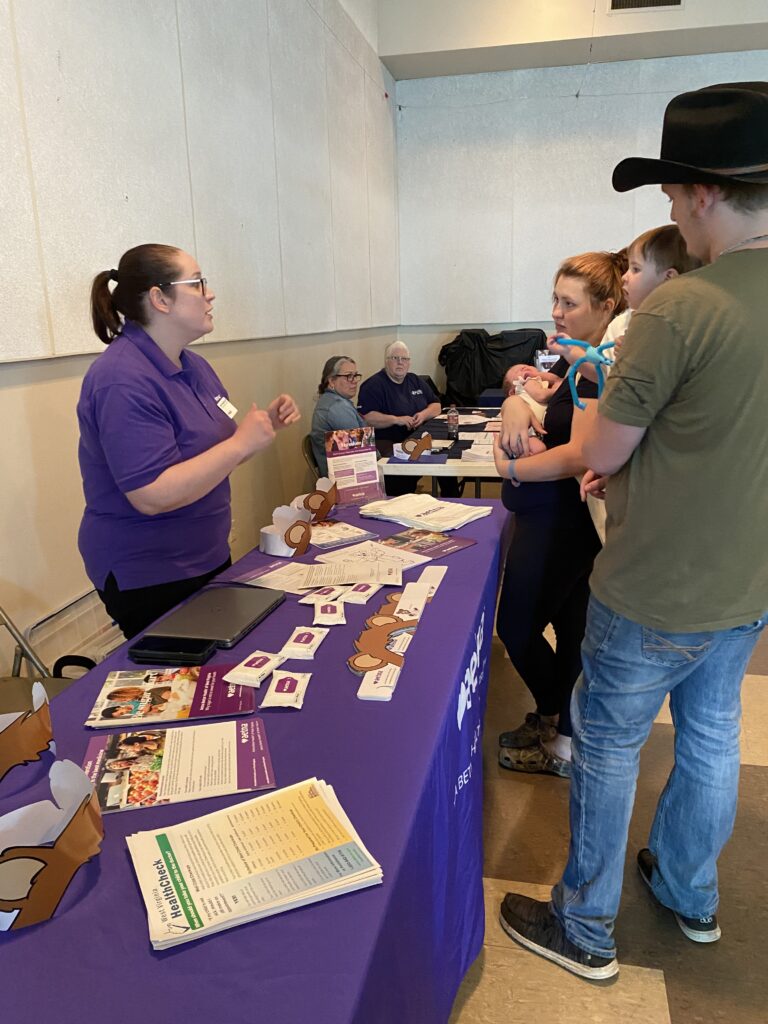 At a purple vendor table, a woman stands in a purple shirt, presenting a couple with paper resources. The woman who is a part of the couple is holding a newborn baby. The man in the cowboy hat holds a toddler.