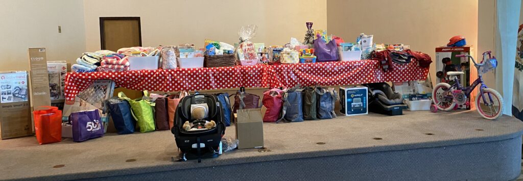 On a long table with a red tablecloth with white polka-dots are a variety of baby items. In front of the table are filled bags and a car seat.