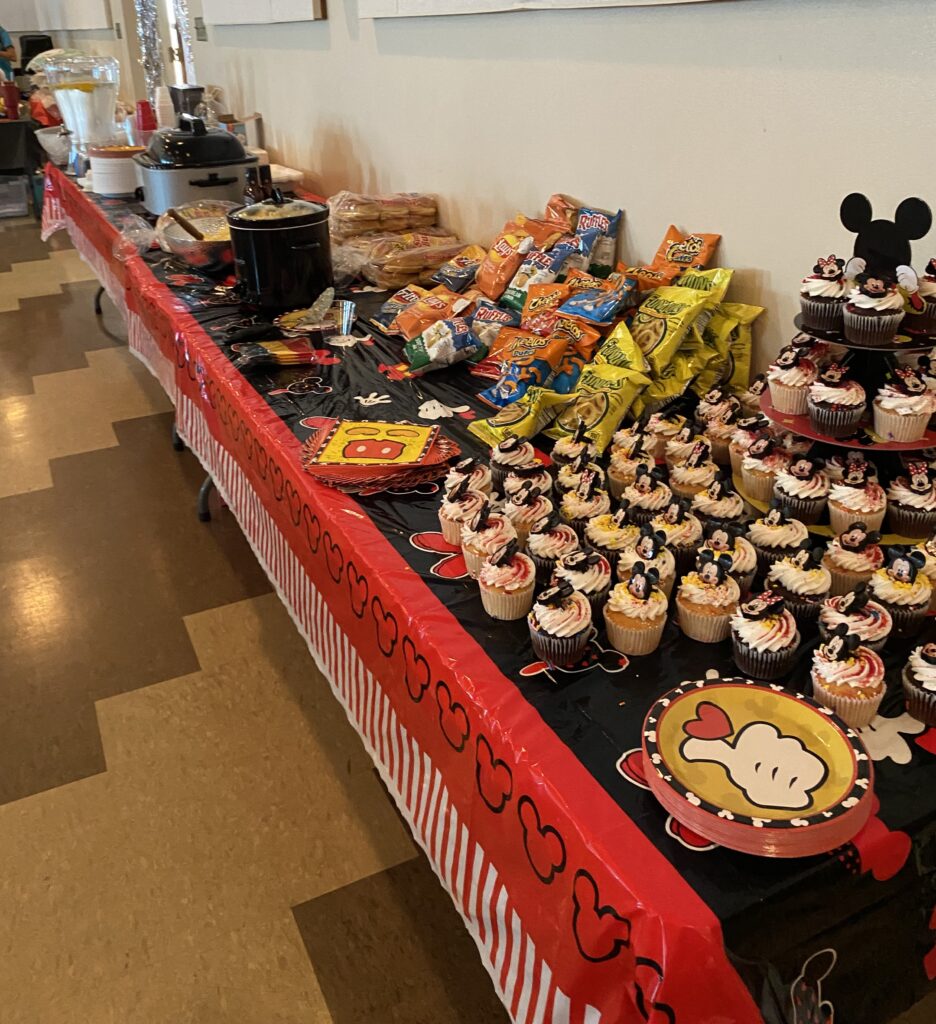 On a black and red table sits a Mikey Mouse cupcake stand, stacked with and surrounded by a variety of chocolate and vanilla cupcakes. Each cupcake has a smiling Mickey Mouse or Minnie Mouse face. As the table stretches on, it is stacked with individual chip bags, slow-cookers, and cups.