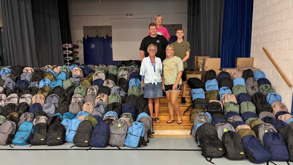 Five people standing on bleachers surrounded by rows of colorful backpacks in a gymnasium, suggesting a school supply drive or community event.