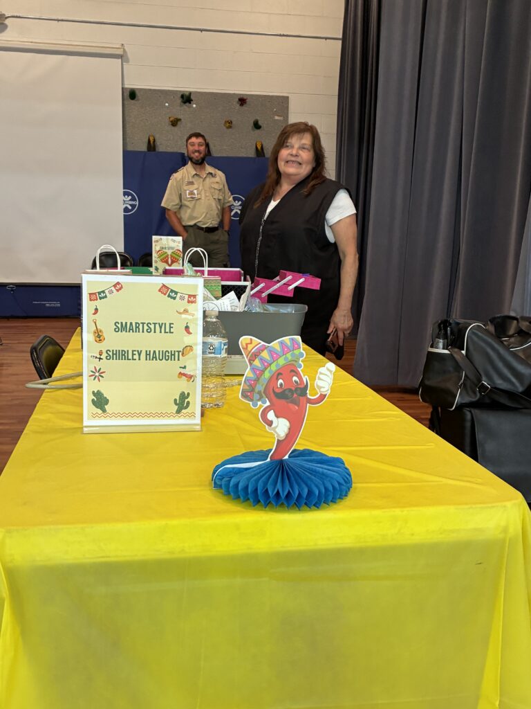 A woman stands in a room near a yellow table covered with colorful decorations. A sign reads "SmartStyle Shirley Haught." A man stand smiling the background. The atmosphere is festive.