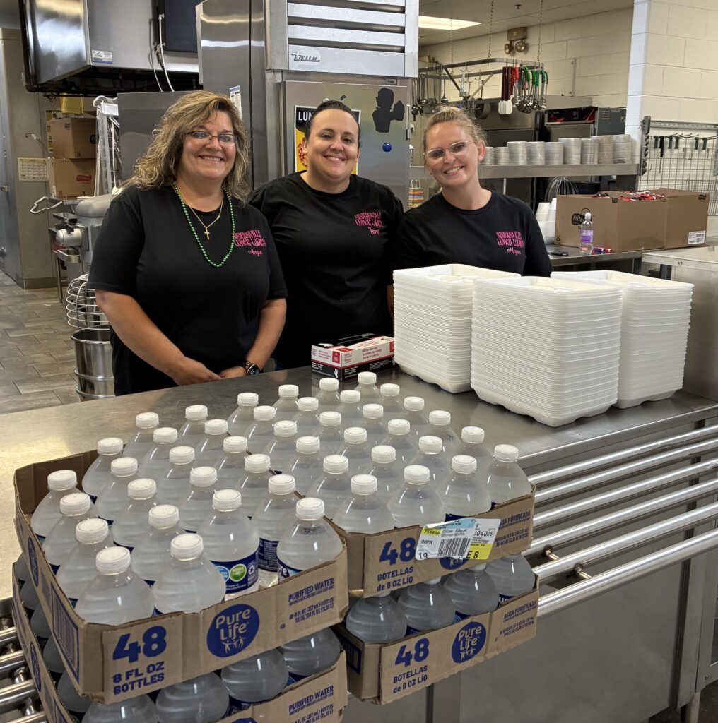 Three smiling women in black shirts stand behind a kitchen counter with stacks of plastic containers and water bottles. Kitchen equipment is visible in the background.