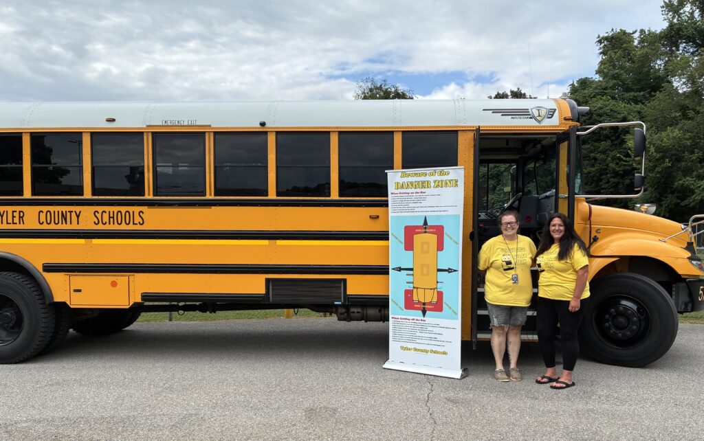 Two people in yellow shirts stand beside a yellow school bus and a sign about a bus safety zone. The scene conveys a sense of safety and community.