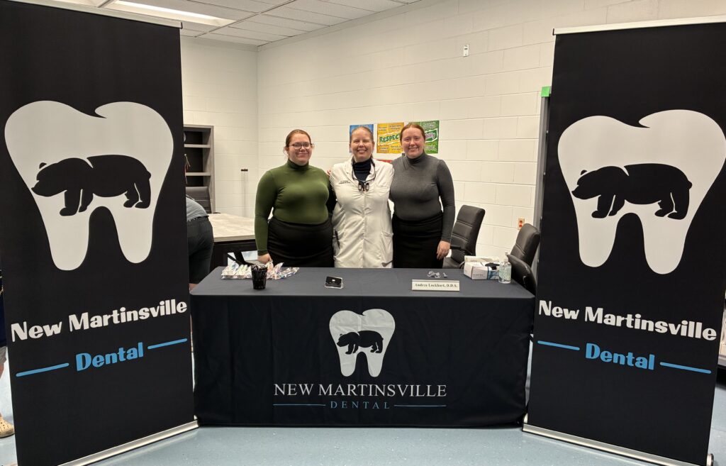 Three people stand smiling behind a table covered in a "New Martinsville Dental" cloth. Two banners with a tooth and bear logo are on each side.