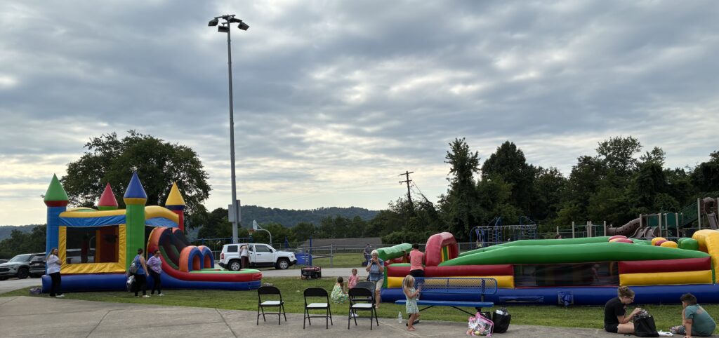 Children play outside on colorful inflatable bounce houses at a park. Chairs are set up nearby. The cloudy sky and trees create a relaxed atmosphere.