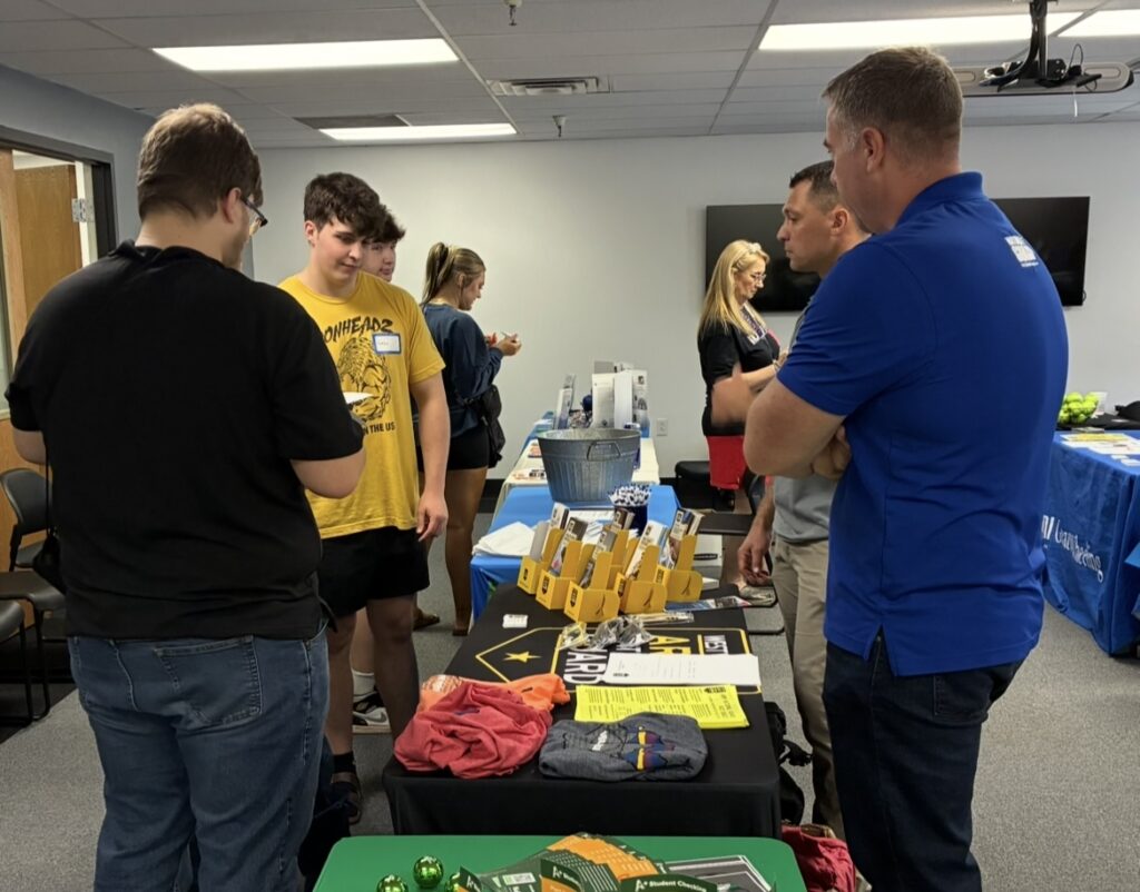 People are gathered around a table at a career fair. The table is filled with brochures and items. The mood is engaging and informative.