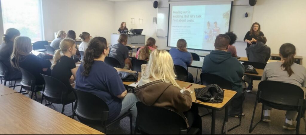 A classroom with students seated at desks facing a projector screen displaying a presentation about moving out and costs. A woman stands near the screen talking.