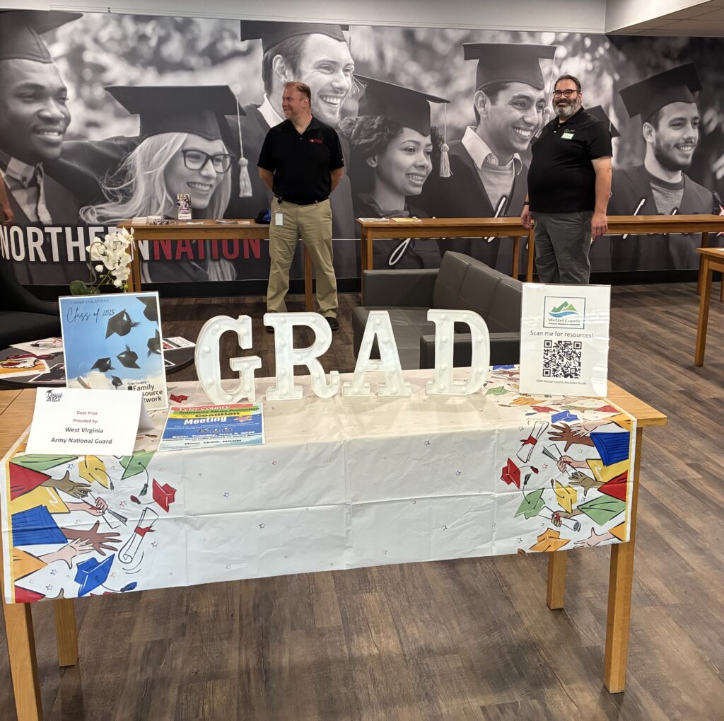 Graduation-themed setup with large "GRAD" letters on a colorful table. Two people stand in front of a mural of smiling graduates in caps.