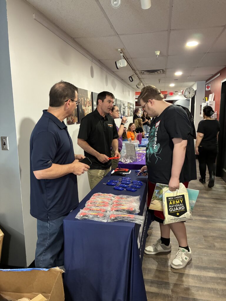 A busy recruitment fair at a hallway with people engaging. Men at a table with promotional items converse with a young man holding a National Guard booklet.