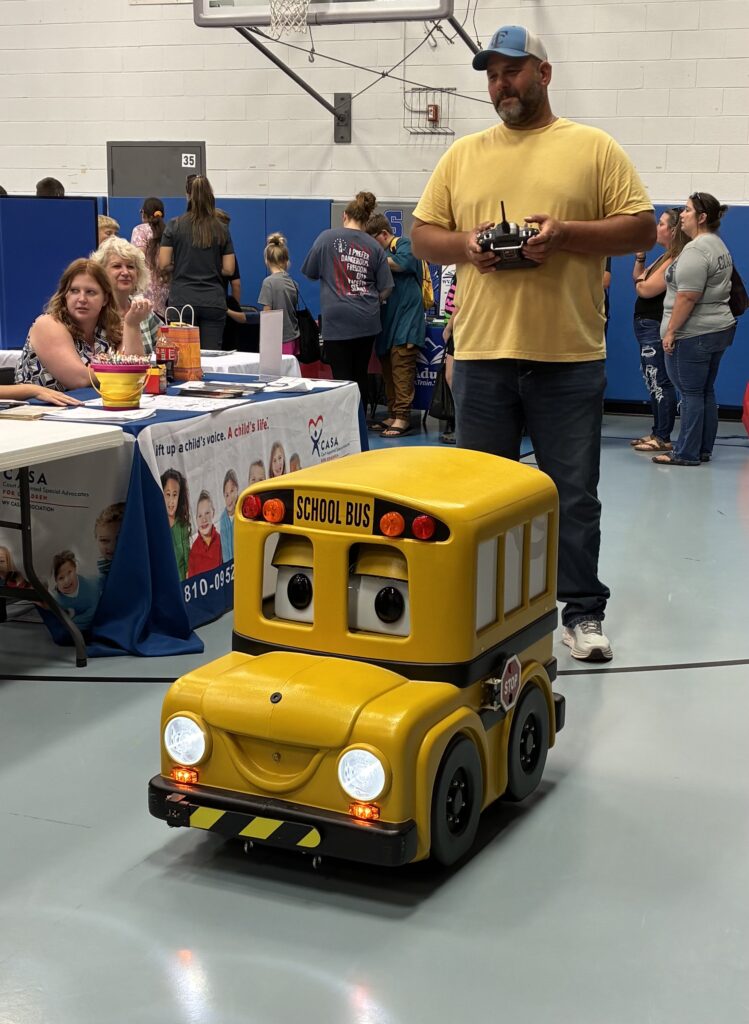 A man operates a remote-controlled, toy-like yellow school bus with animated eyes in a bustling indoor event. People and informational tables are in the background.