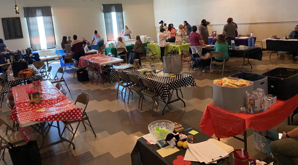 A community room with black and white chevron floors holds many folding tables with red and white or black and white polka-dotted tablecloths. In the back of the room stands a group of people.