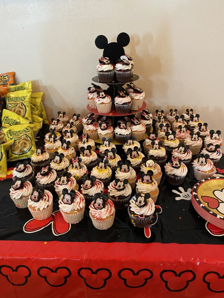 On a black and red table sits a Mikey Mouse cupcake stand, stacked with and surrounded by a variety of chocolate and vanilla cupcakes. Each cupcake has a smiling Mickey Mouse or Minnie Mouse face.