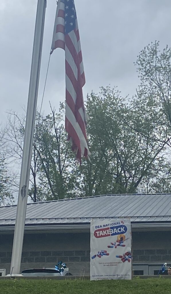 American flag flying at half-mast beside a building with a DEA National Take Back banner. Overcast sky and trees are visible in the background.