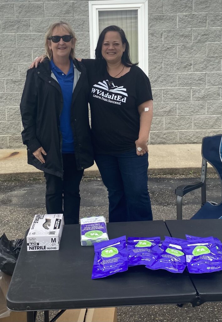 Two people smiling outdoors, one wearing sunglasses, standing by a table with safety gloves and purple packs. The mood is friendly and welcoming.