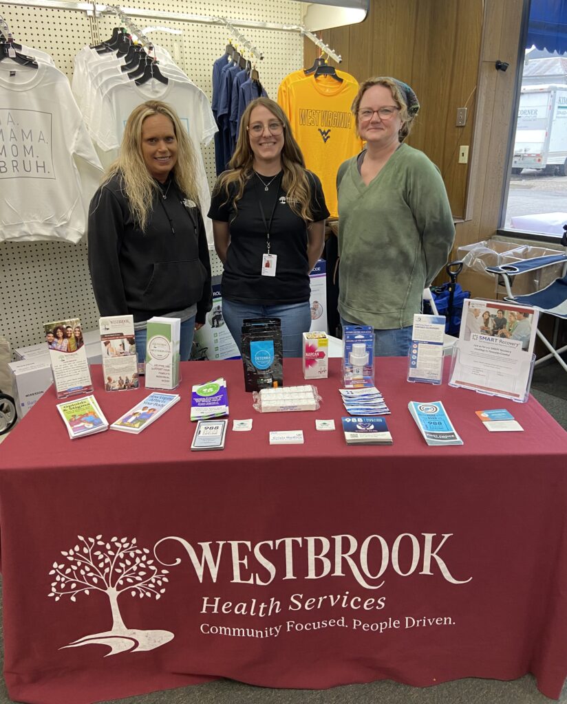 Three people stand smiling behind a table covered with brochures and products. The tablecloth reads "Westbrook Health Services." Clothes hang in the background.