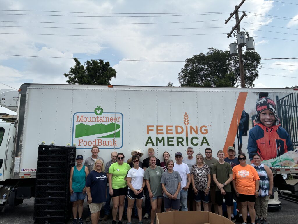 Group of volunteers smiling in front of a Mountaineer Food Bank and Feeding America truck. The scene conveys a sense of community and teamwork.