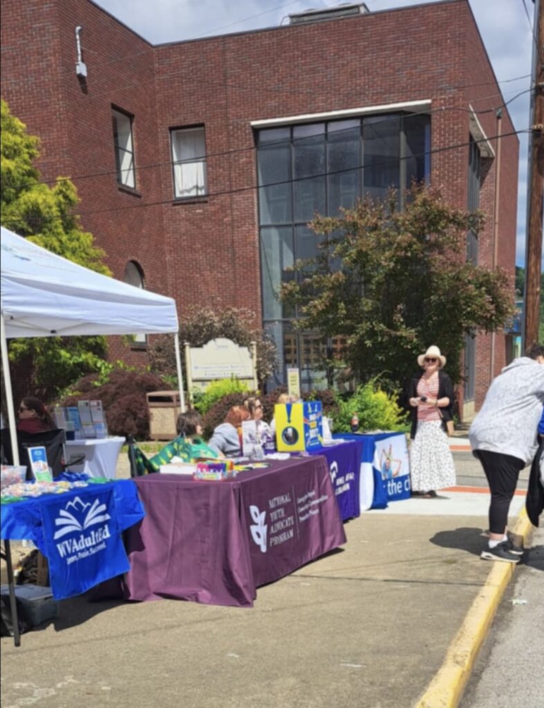 Outdoor community event with tables displaying informational banners. People browse in sunny weather beside a brick building. Atmosphere is lively and engaging.