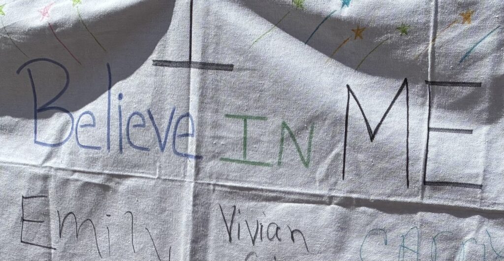 A white cloth with colorful text reading "Believe in Me" surrounded by various hand-drawn names and star doodles, conveying a hopeful and supportive tone.