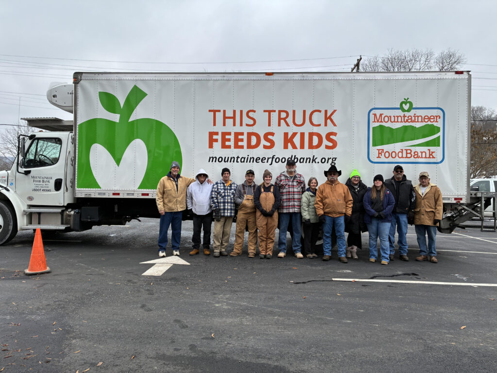 A group of people in winter clothing stand smiling in front of a large truck. The truck's side reads, "This Truck Feeds Kids," promoting Mountaineer Food Bank.