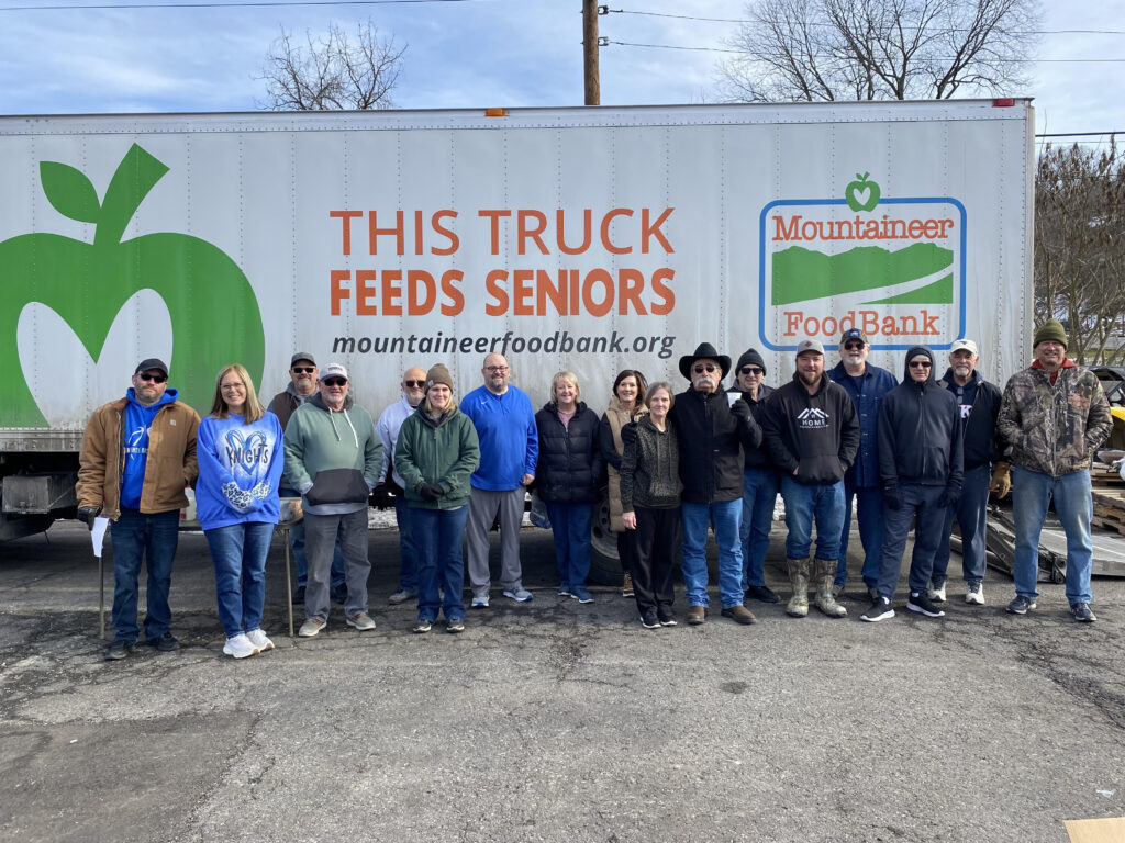 A group of 16 people stand smiling in front of a truck with "This Truck Feeds Seniors" and "Mountaineer Food Bank" logos. They appear cheerful and united.