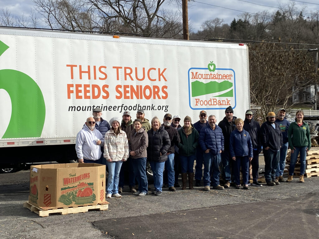 A group of people stand smiling in front of a large truck labeled "This Truck Feeds Seniors" and "Mountaineer FoodBank." A box of watermelons is nearby.