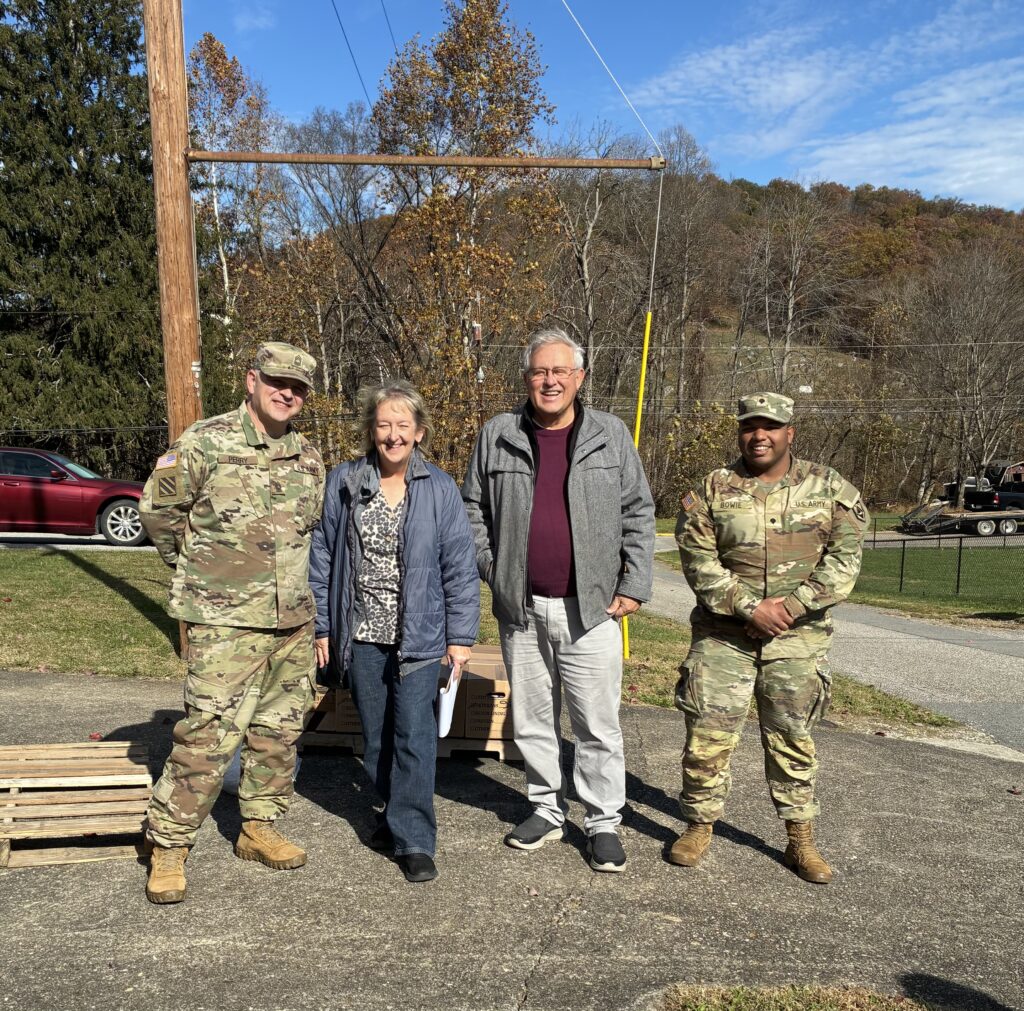 Two men in military uniforms and a casually dressed man and woman stand smiling outdoors on a sunny day, with autumn trees in the background.