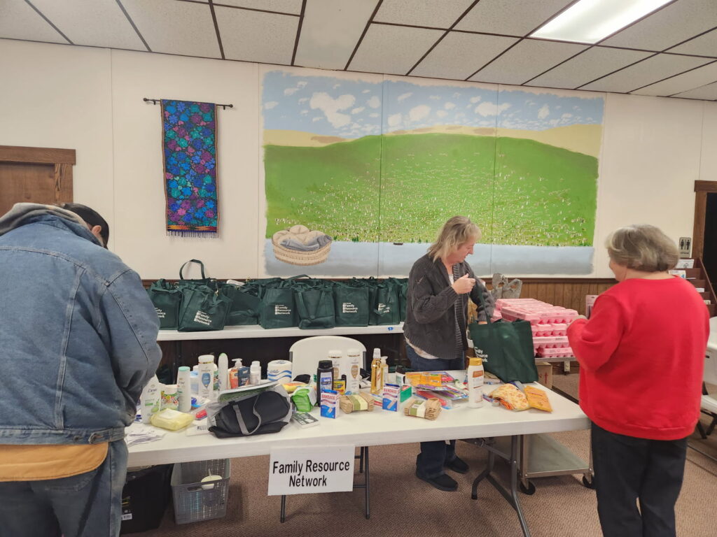 Three people organize supplies on a table labeled "Family Resource Network." Bags and household items are present. A mural of a green landscape decorates the wall.