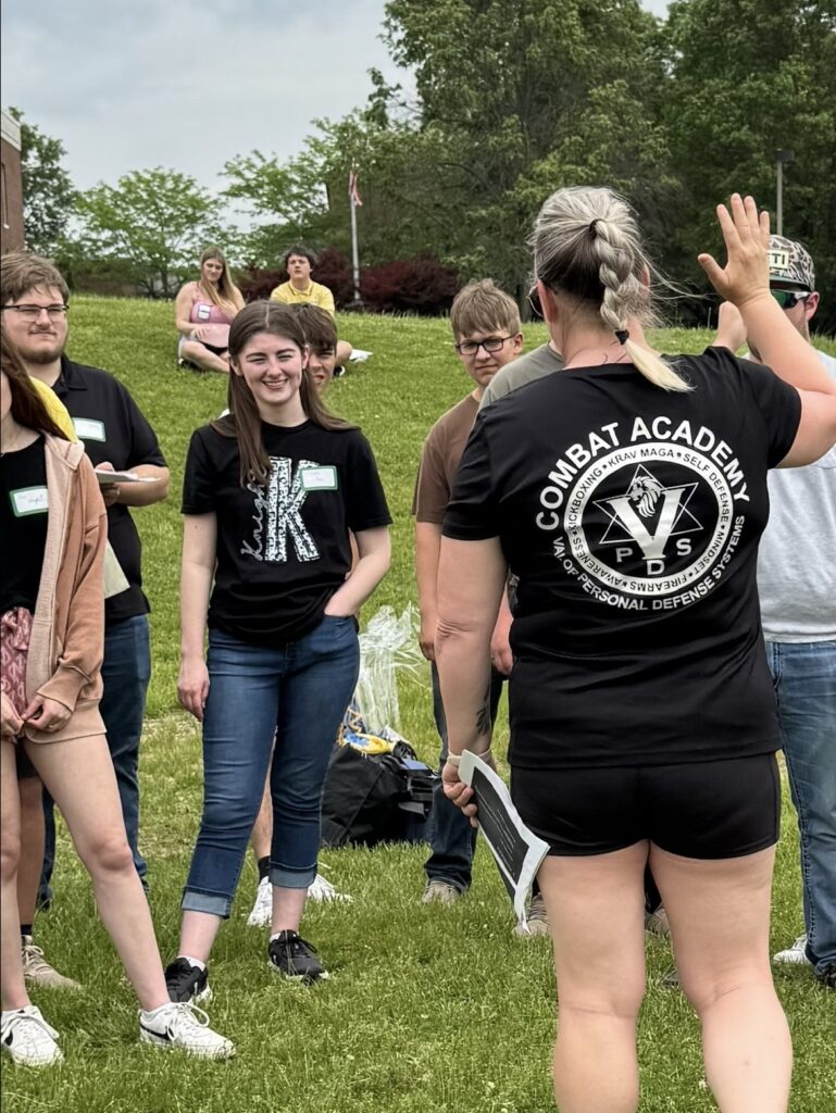 A woman in a "Combat Academy" shirt instructs a group of smiling young adults outdoors on grass, creating a lively, engaging atmosphere.