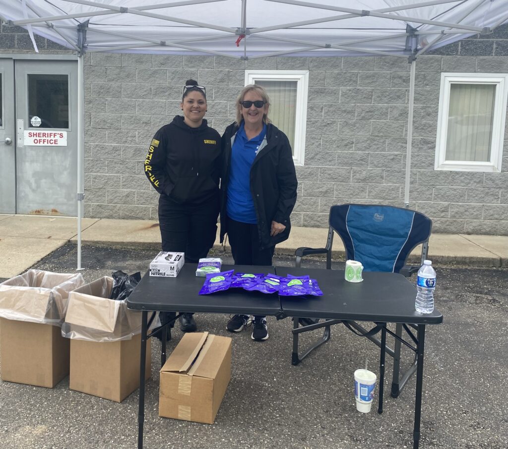 Two people smiling under a canopy in front of a building labeled "Sheriff's Office." A table has packaged items, a water bottle, and empty boxes.